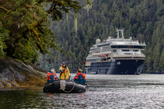 Ein Schlauchboot mit 3 Personen im Vordergrund, im Hintergrund ein Expeditionsschiff in einem Fjord